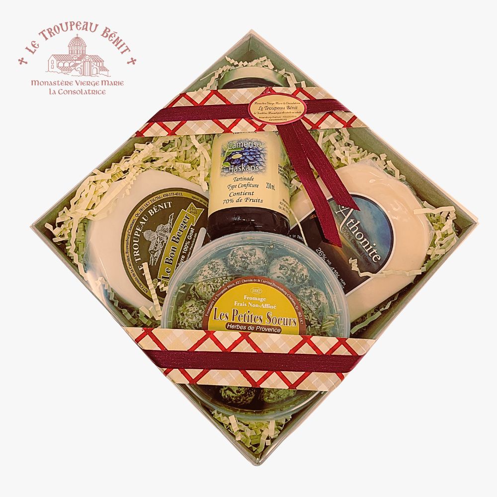 A gift box of local made cheeses and from Le Troupeau Benit, arranged in a diamond orientation on a white background. The box contains several round and oval cheeses wrapped in white paper, a jar of jam, and a round container of fresh goat cheese balls, all nestled in white shredded paper and tied with red patterned ribbons.